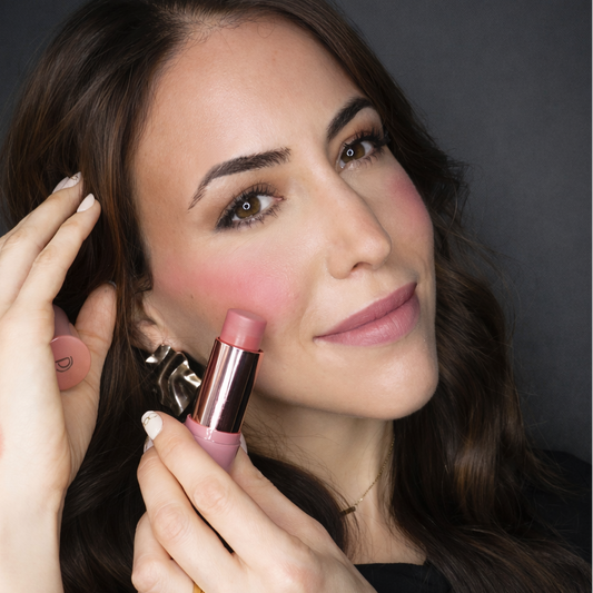 Woman applying blush to her cheeks with a pink makeup stick against a dark background
